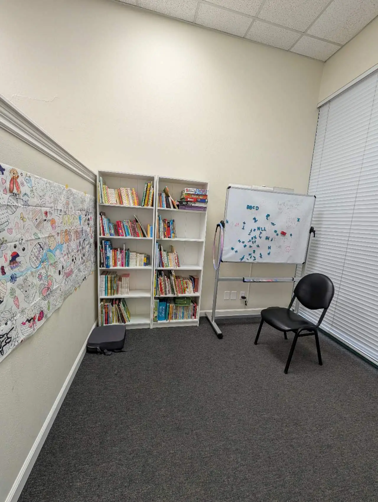 A corner of the library: two white bookcases full of children’s books, a whiteboard, and a wall of children’s coloring pages