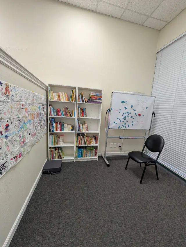 A corner of the library: two white bookcases full of children's books, a whiteboard, and a wall of children's coloring pages